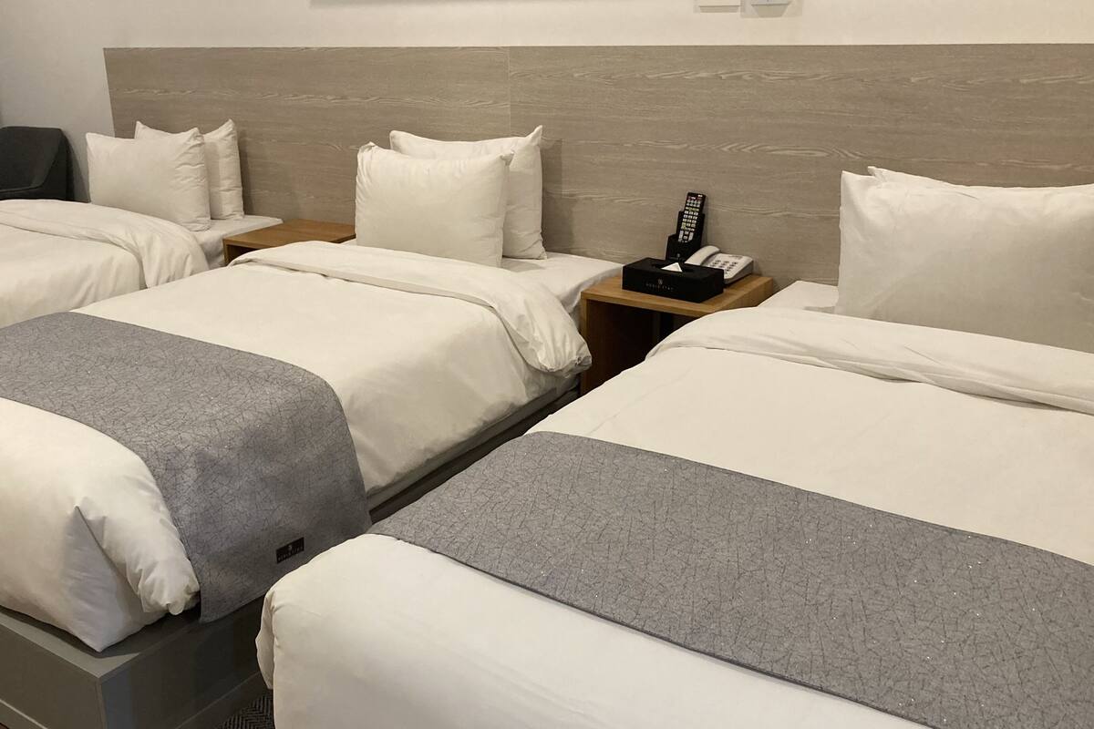 Habitación de hotel con dos camas, cada una con ropa blanca y detalles grises. Tiene un cabecero de madera, una mesita de noche con un teléfono y una caja, y una silla en la esquina.