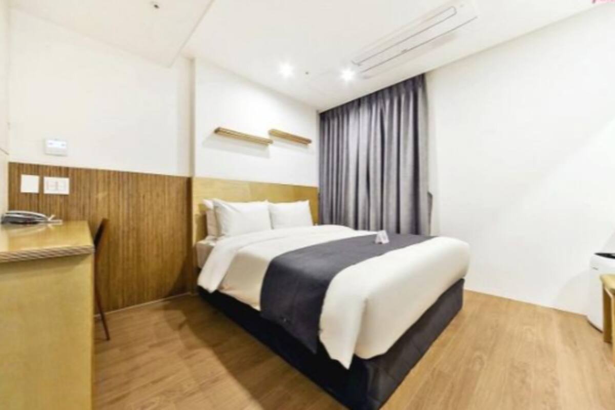 Habitación de hotel con cabecera de madera, cama con ropa de cama blanca y oscura, escritorio con teléfono y ventana con cortinas.