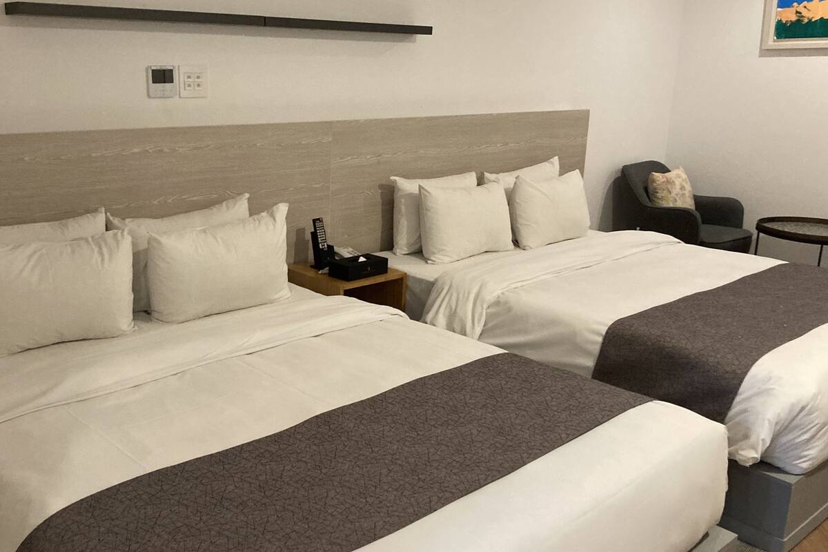 Habitación de hotel con dos camas, una mesita de noche con control remoto, una silla y una pequeña mesa.