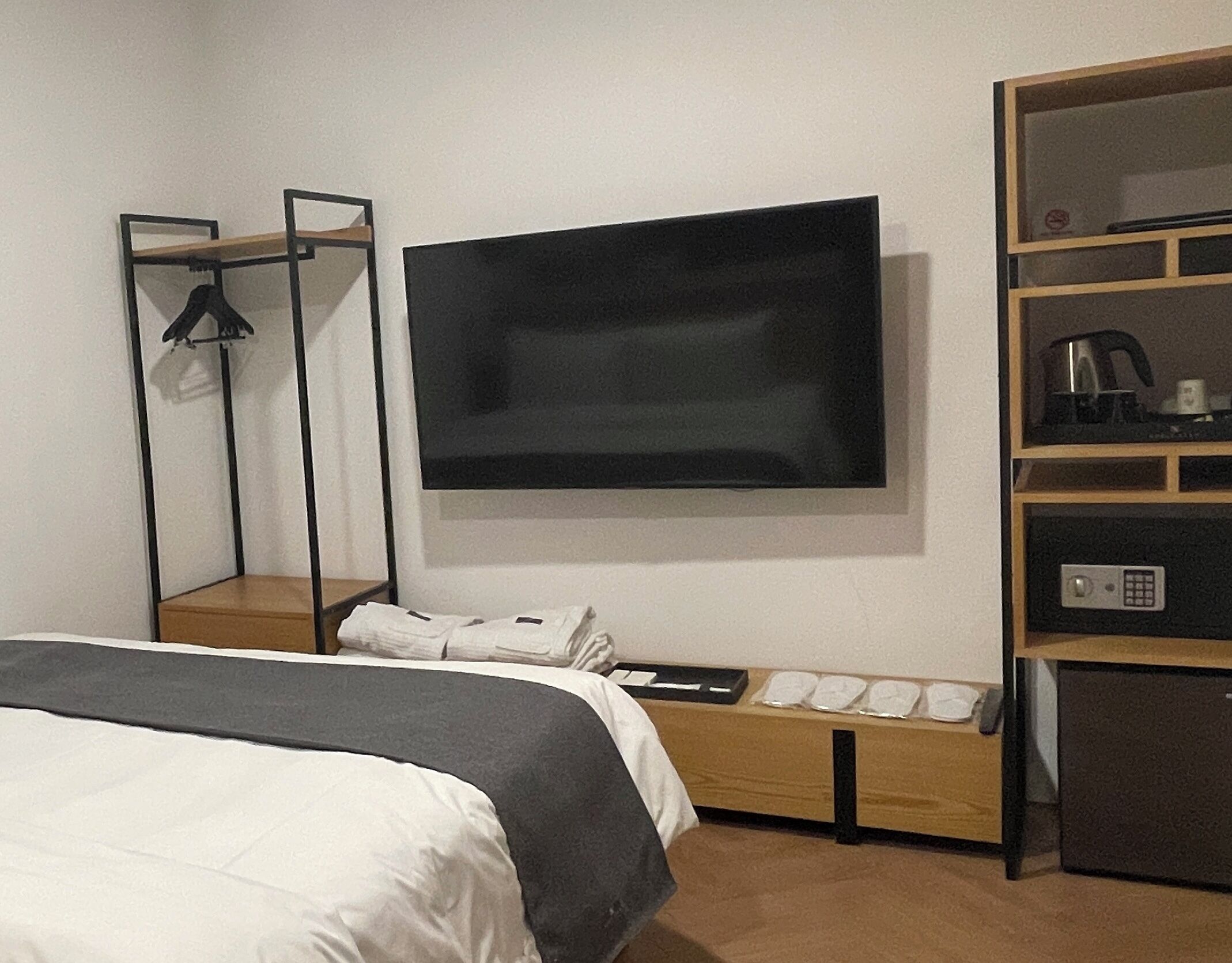 A modern hotel room with a large bed, a flat-screen TV mounted on the wall, a wooden bedside table with a remote control, and a built-in shelf with a kettle and other items.