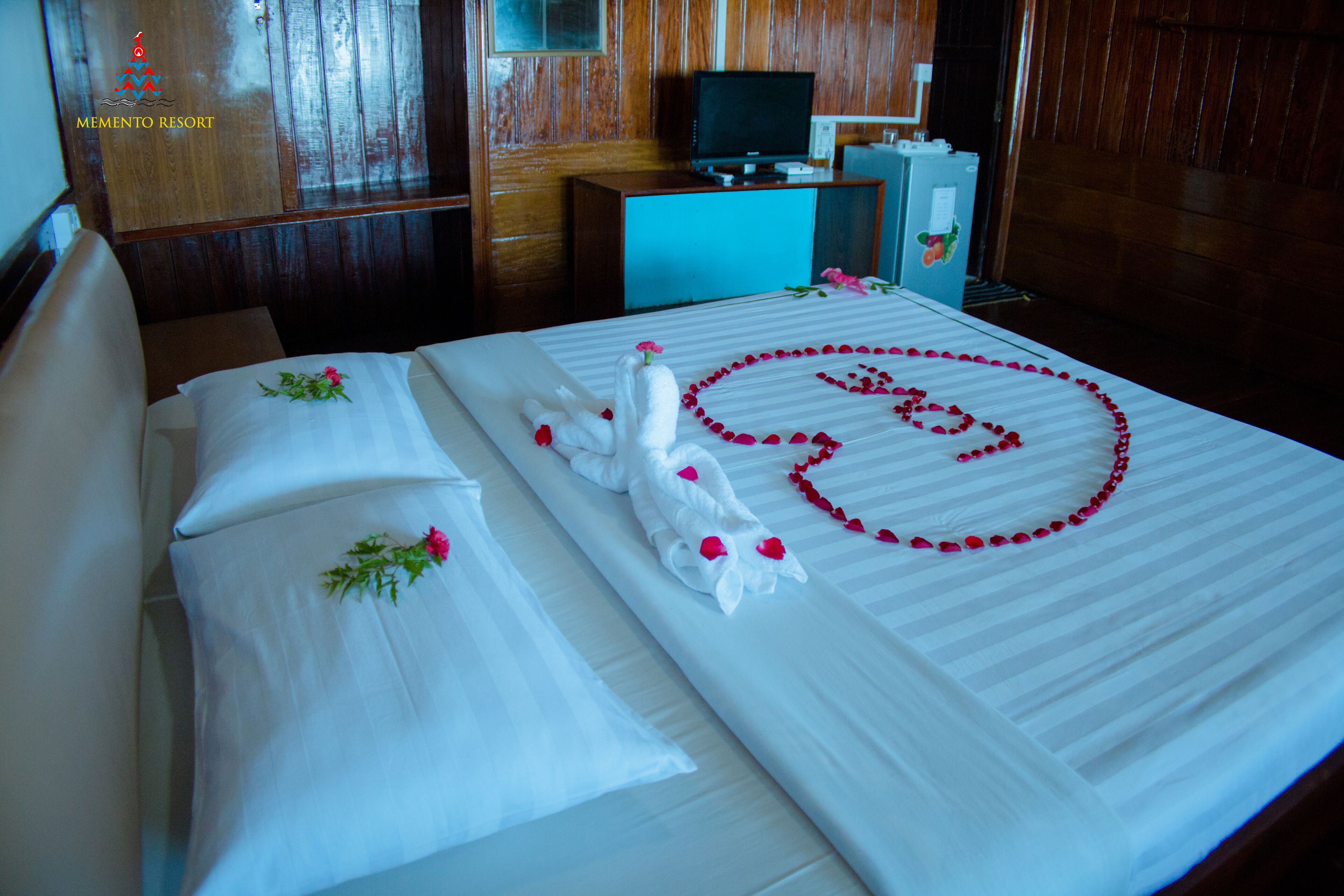 A neatly made bed with a heart-shaped arrangement of rose petals, a white towel, and a decorative swan.