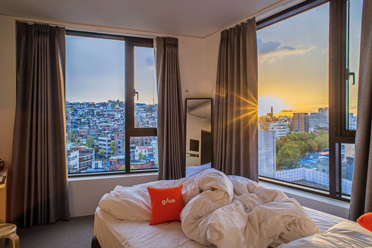 A hotel room with a large window offering a city view, a bed with white bedding and an orange pillow, and a desk with a chair.