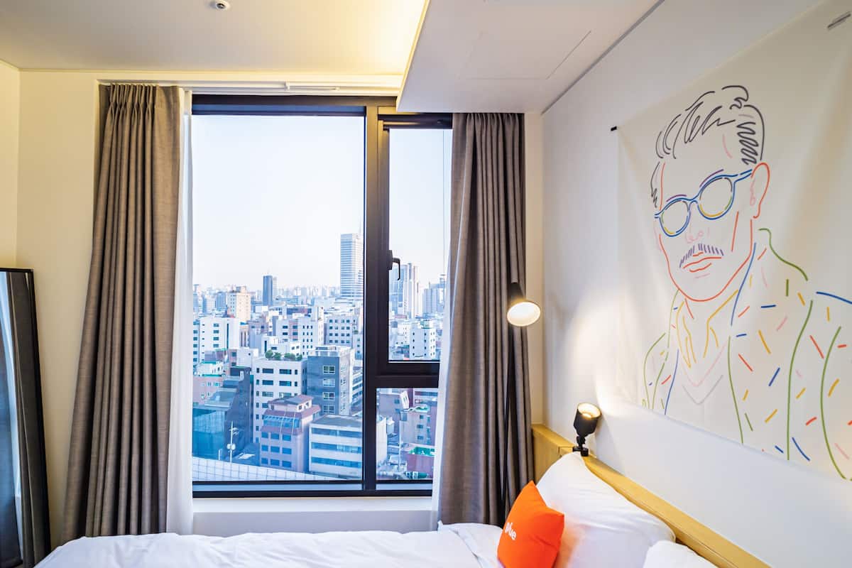 A hotel room with a large window, a bed with white bedding, an orange pillow, and a wall with a colorful abstract painting.