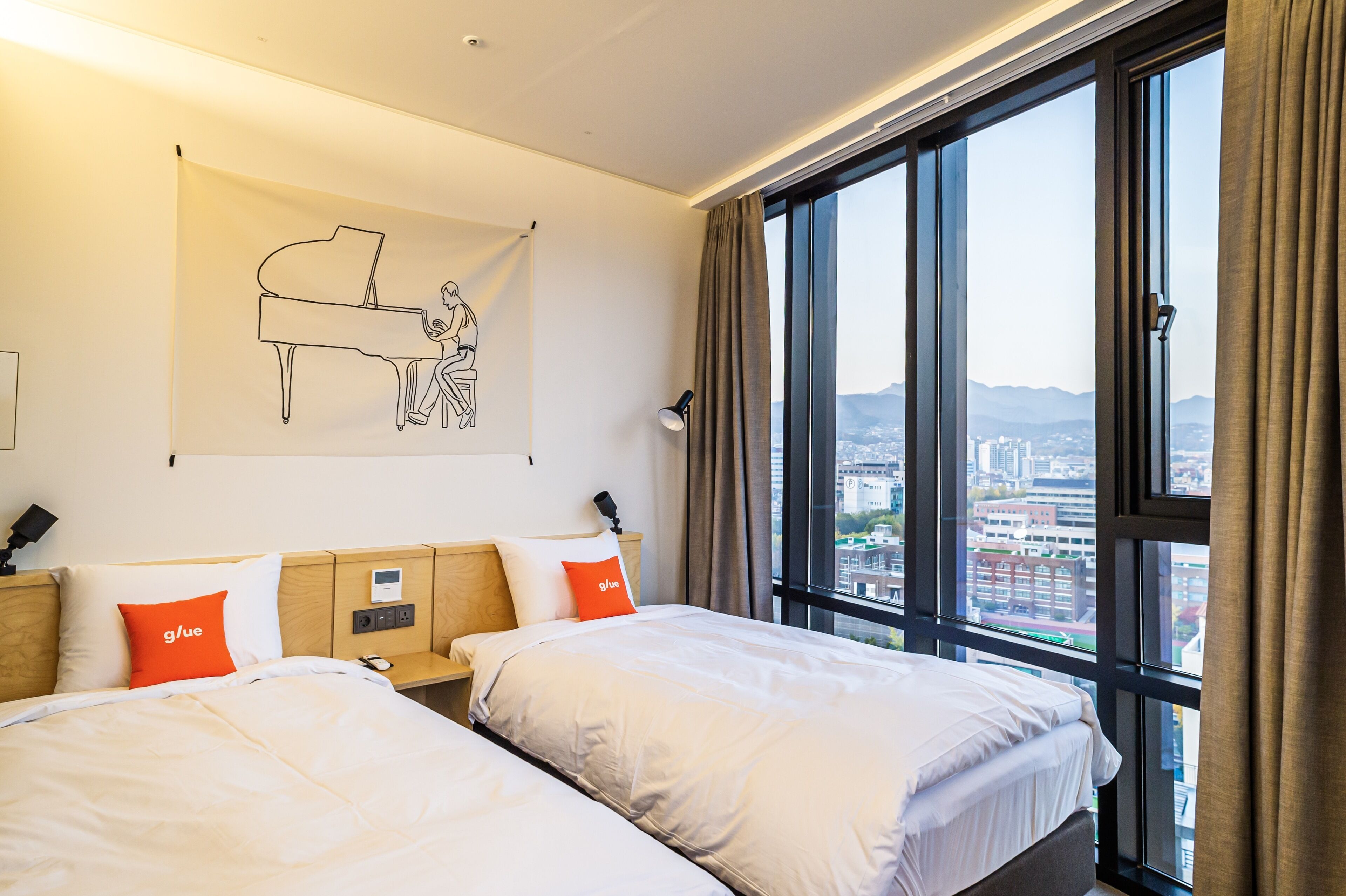 Une chambre d’hôtel avec deux lits, une illustration de piano sur le mur, une vue sur la ville à travers de grandes fenêtres et un panneau de commande sur la table de chevet.