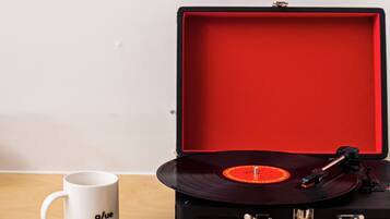 A vinyl record player in a red case, a white mug with "g/ue" on it, and a framed poster on the wall.