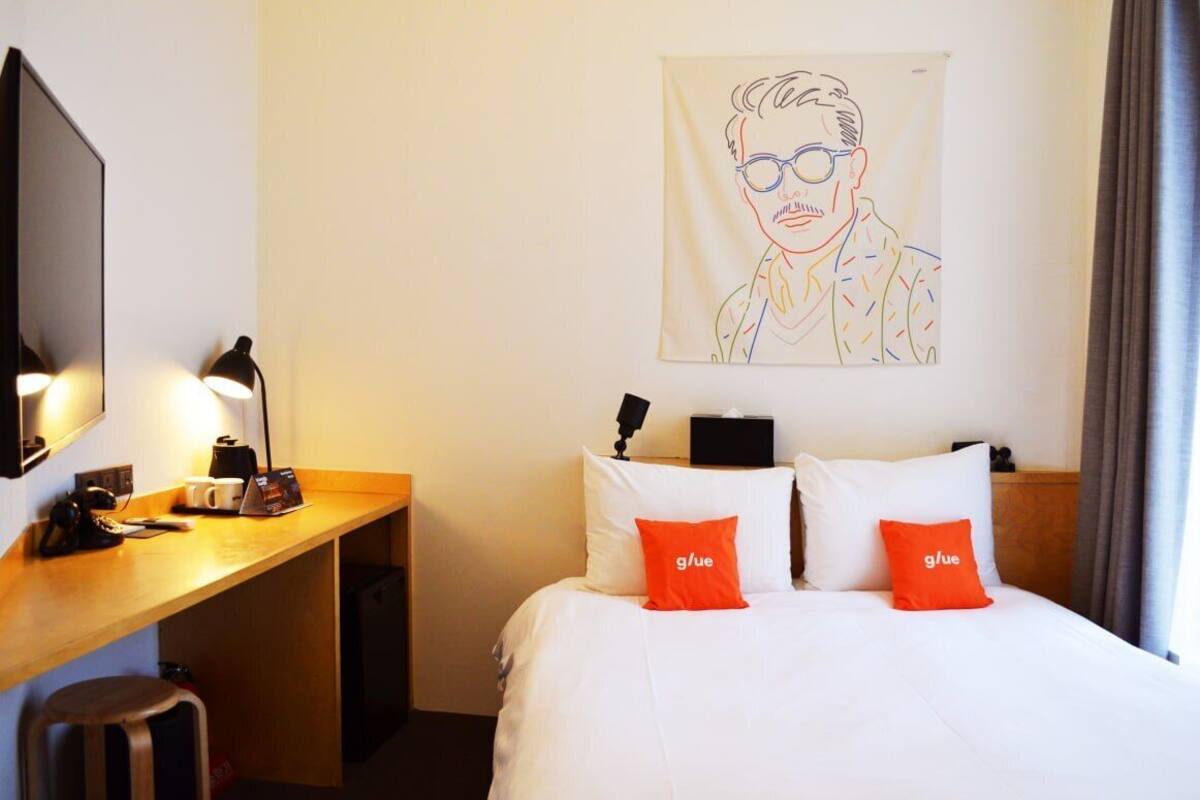A hotel room with a bed, a desk with a computer, a lamp, and a wall-mounted artwork.