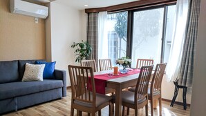 A modern living room with a grey sofa, wooden dining table, and chairs. There is a potted plant and a vase with flowers on the table. - Grato Terrace (Tokyo)