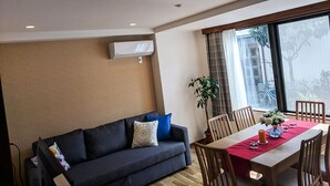 A modern living room with a dark sofa, a dining table with chairs, and a large window with a view of buildings and trees. - Grato Terrace (Tokyo)