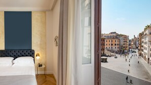 Habitación de hotel con una cama grande, una mesita de noche con lámpara y vistas a una calle concurrida y edificios históricos.