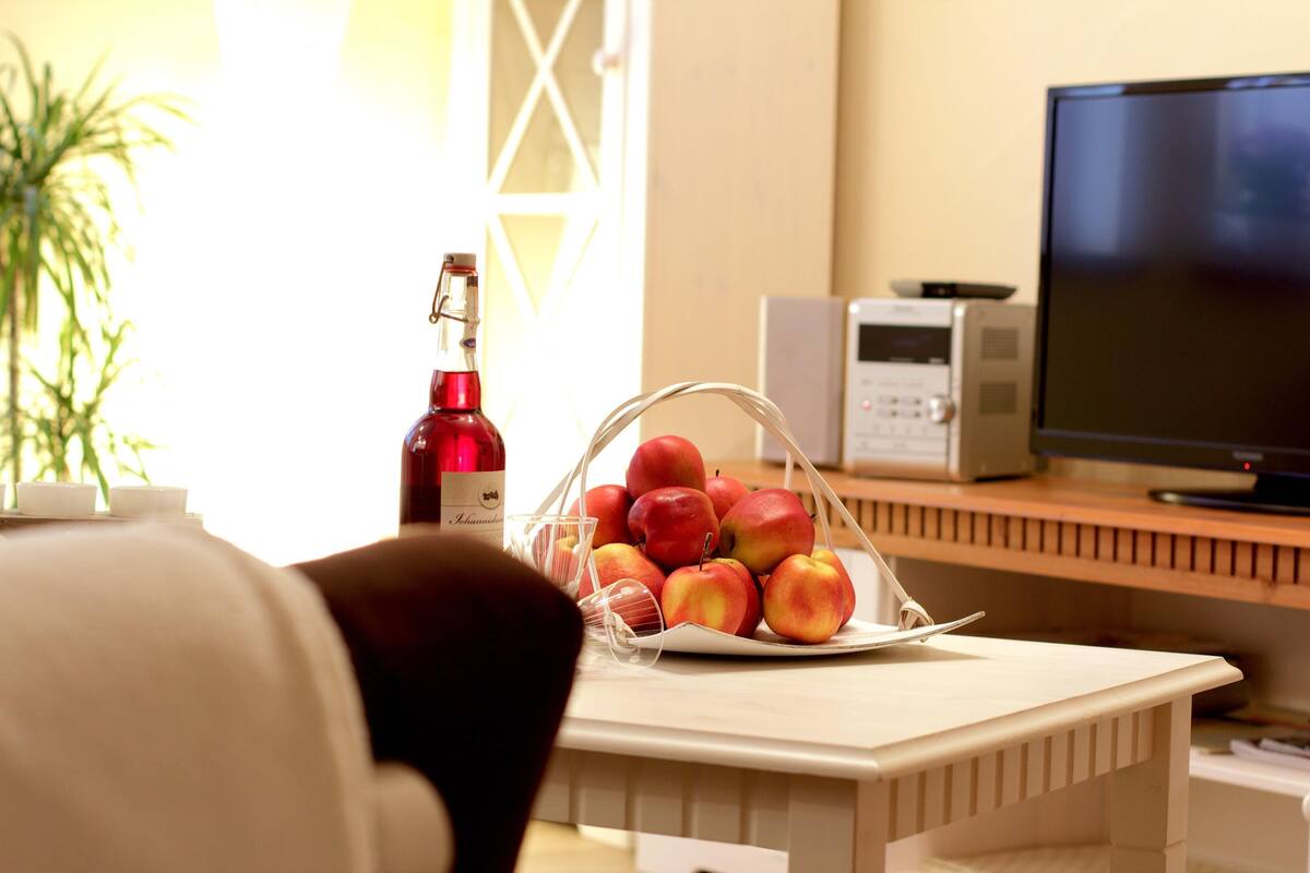 A room with a wooden table, a basket of apples, a bottle of wine, and a television.