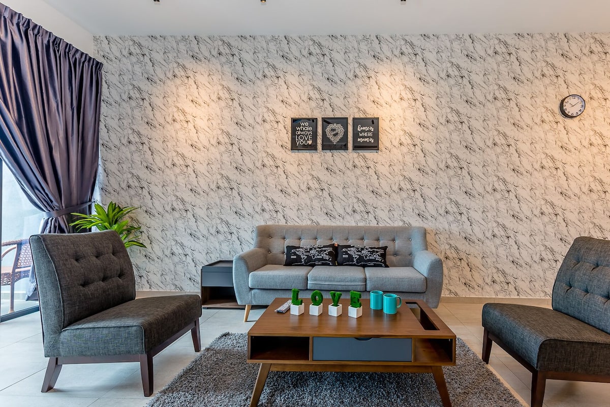 Un salon avec un canapé gris, deux fauteuils, une table basse et un mur orné d’un motif imitant le marbre. Trois tableaux encadrés sont accrochés au mur, ainsi qu’une horloge.
