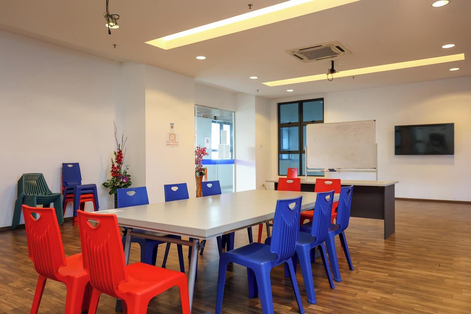 Uma sala de conferências com uma mesa comprida, cadeiras azuis e vermelhas, um quadro branco e uma tela de televisão.