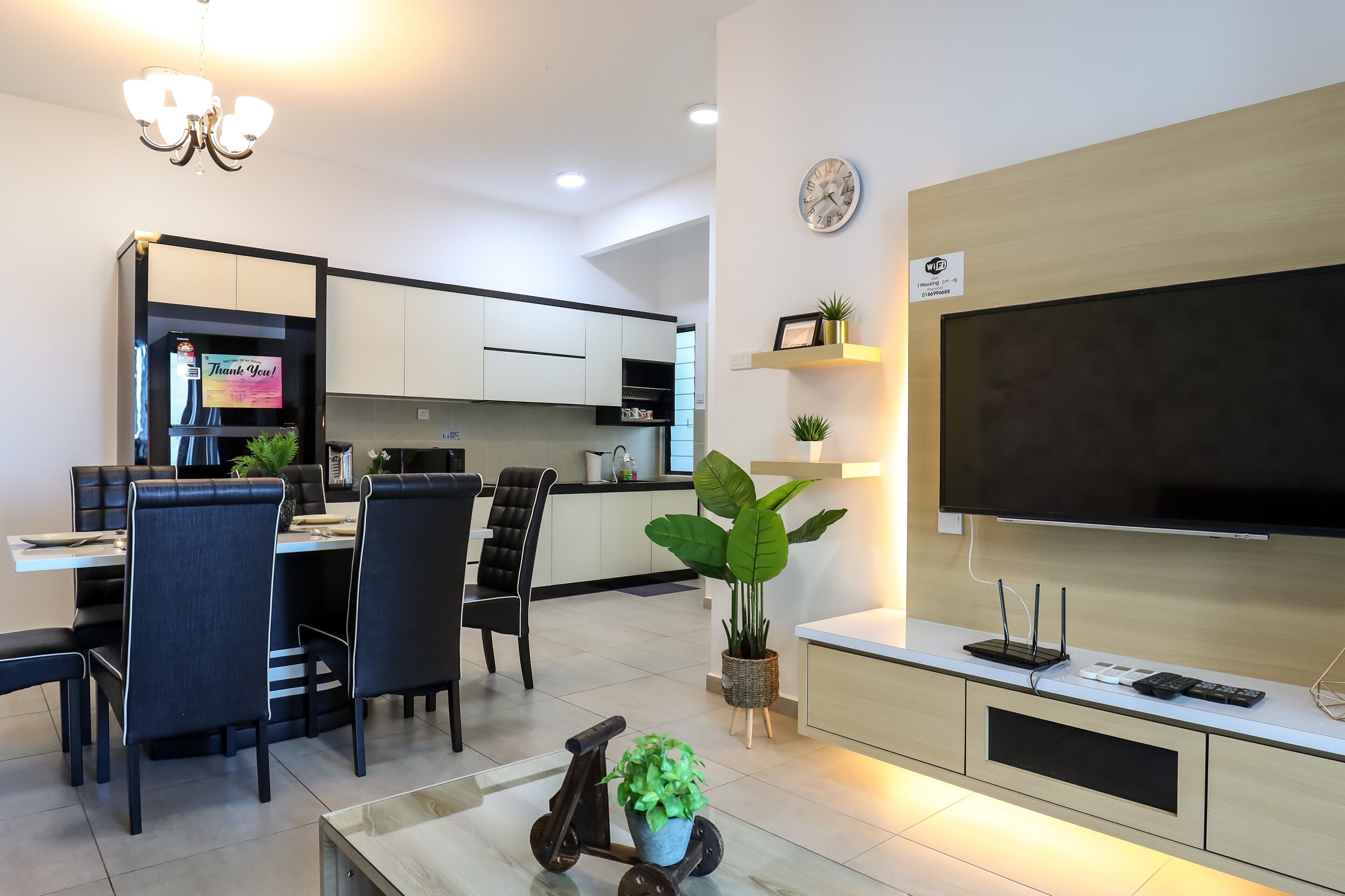 Una sala moderna con un televisor de pantalla plana, una zona de comedor con sillas y una mesa, una cocina al fondo y una lámpara en el techo.