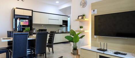 Una sala moderna con un televisor de pantalla plana, una zona de comedor con sillas y una mesa, una cocina al fondo y una lámpara en el techo.