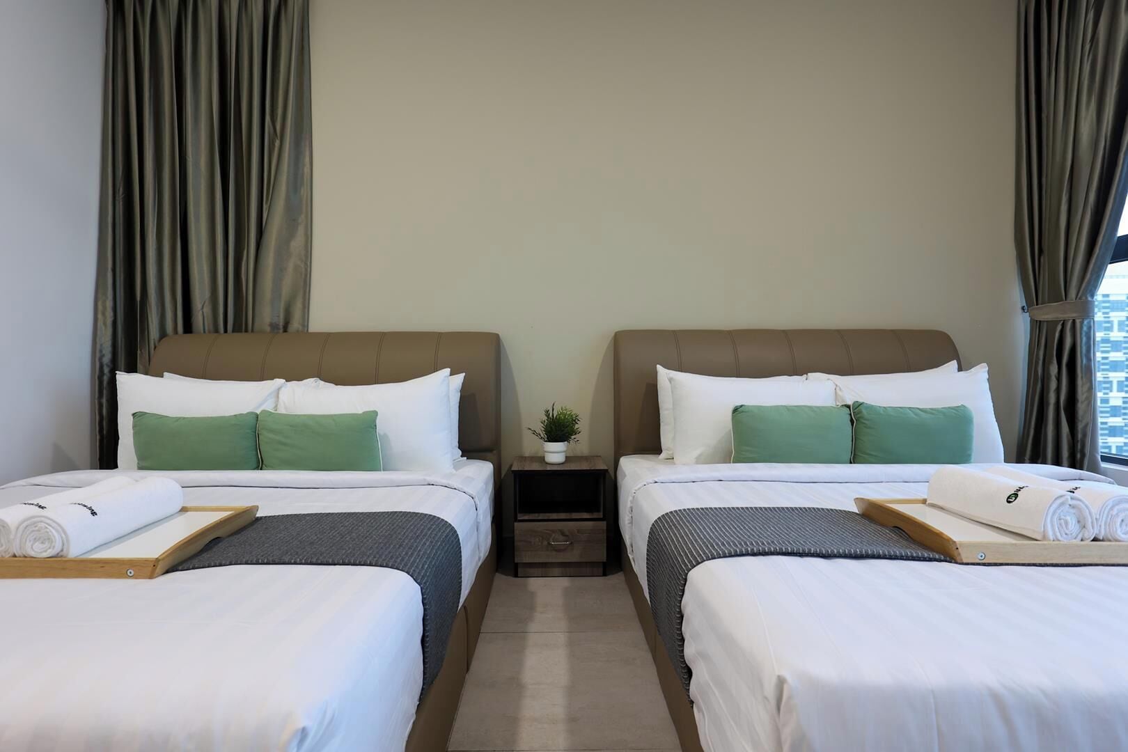 A hotel room with two beds, each with white bedding and green pillows, a small bedside table with a plant, and a view of the city through the window.