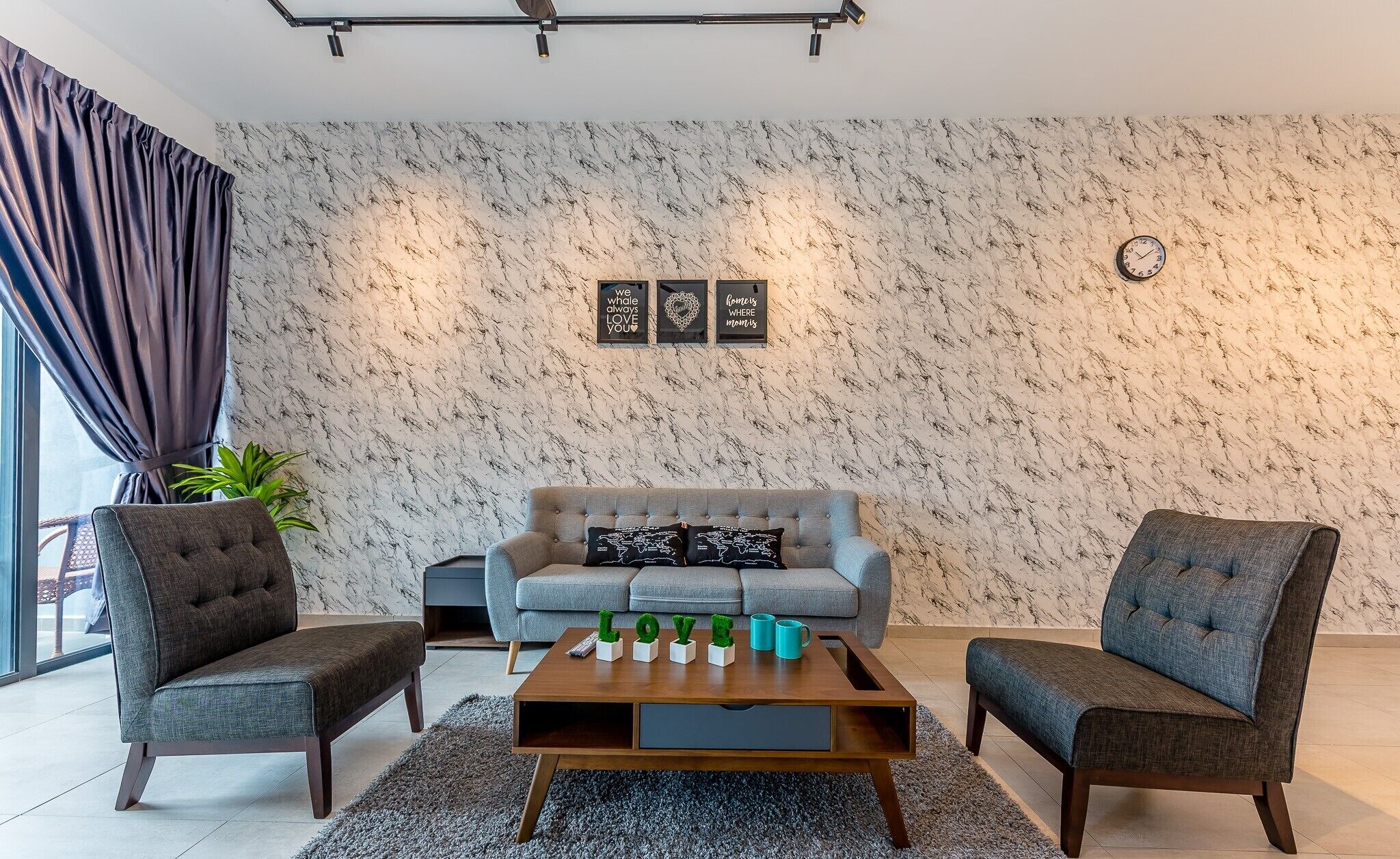 Un salón moderno con pared de diseño de mármol, un sofá gris, dos sillones, una mesa de centro con objetos decorativos y un reloj en la pared.