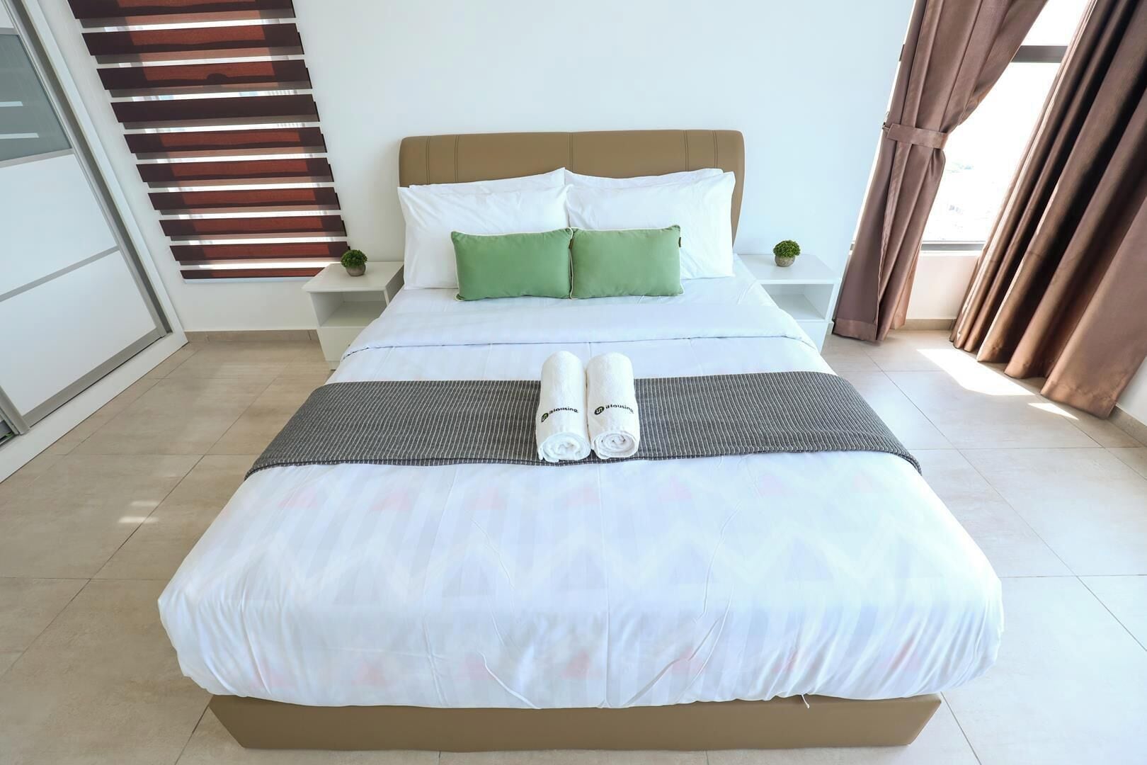 A neatly made bed with a grey bedspread, two rolled towels, and a green pillow.
