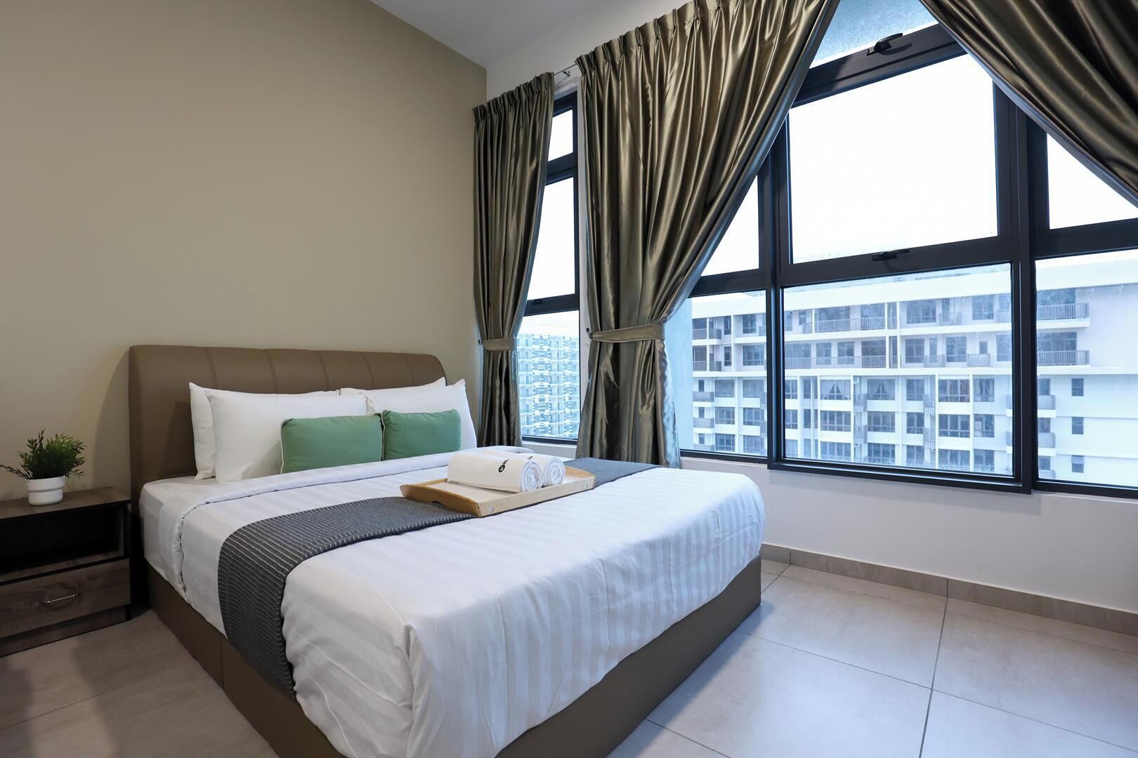 Ein Hotelzimmer mit einem großen Bett, einem Nachttisch mit Pflanze und Blick auf die Stadt durch große Fenster.