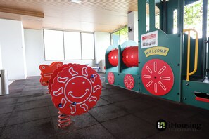 Ein Kinderspielbereich mit einer großen, roten Skulptur im Smiley-Design, einem Willkommensschild und Fitnessgeräten.