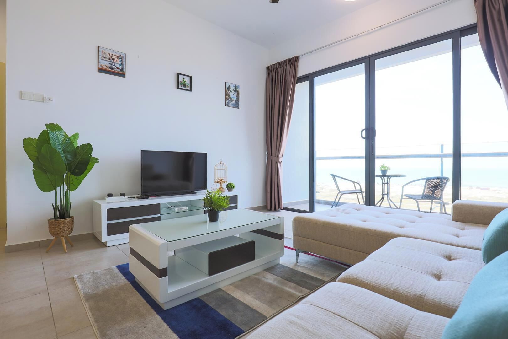 Un soggiorno moderno con televisione a schermo piatto, un tavolino da caffè in vetro, una pianta in vaso di grandi dimensioni, un divano e un balcone con vista sul mare.