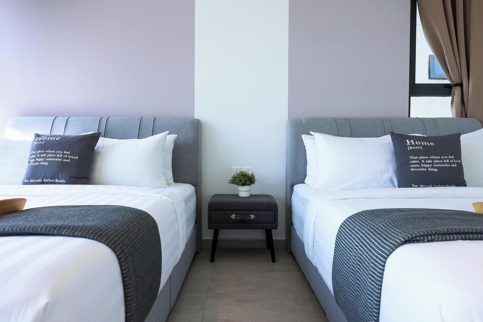 Habitación de hotel con dos camas, cada una con un cojín que dice 'Home [hom]'. Hay una mesita de noche pequeña con una planta entre las camas.