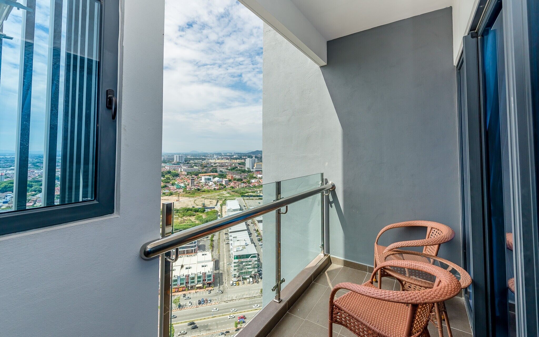 A balcony with a view of a cityscape, a glass railing, and a wicker chair.