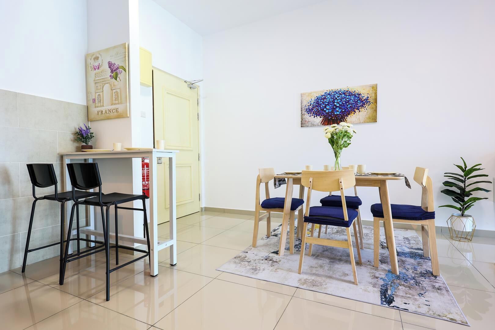 A dining area with a table and chairs, a small plant, and a painting on the wall.