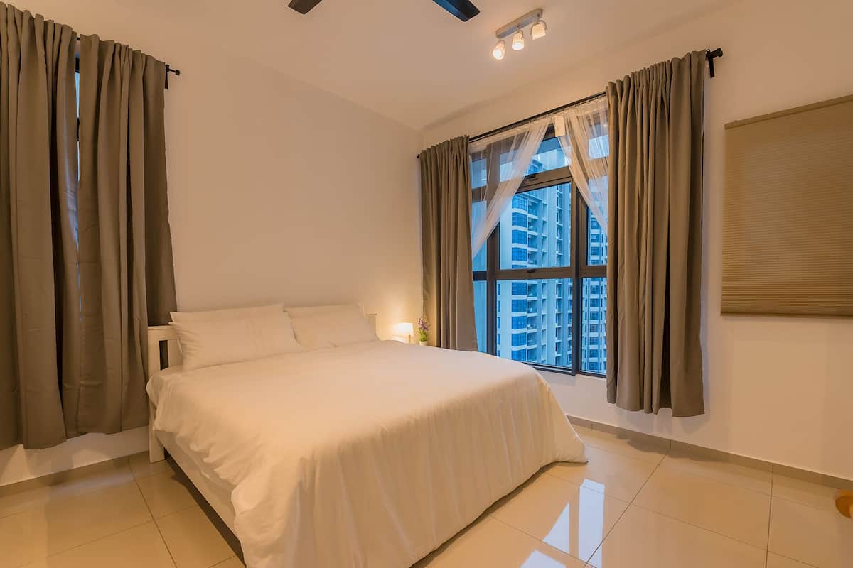 Une chambre à coucher avec un lit, des rideaux, une fenêtre donnant sur des immeubles et un luminaire au plafond.