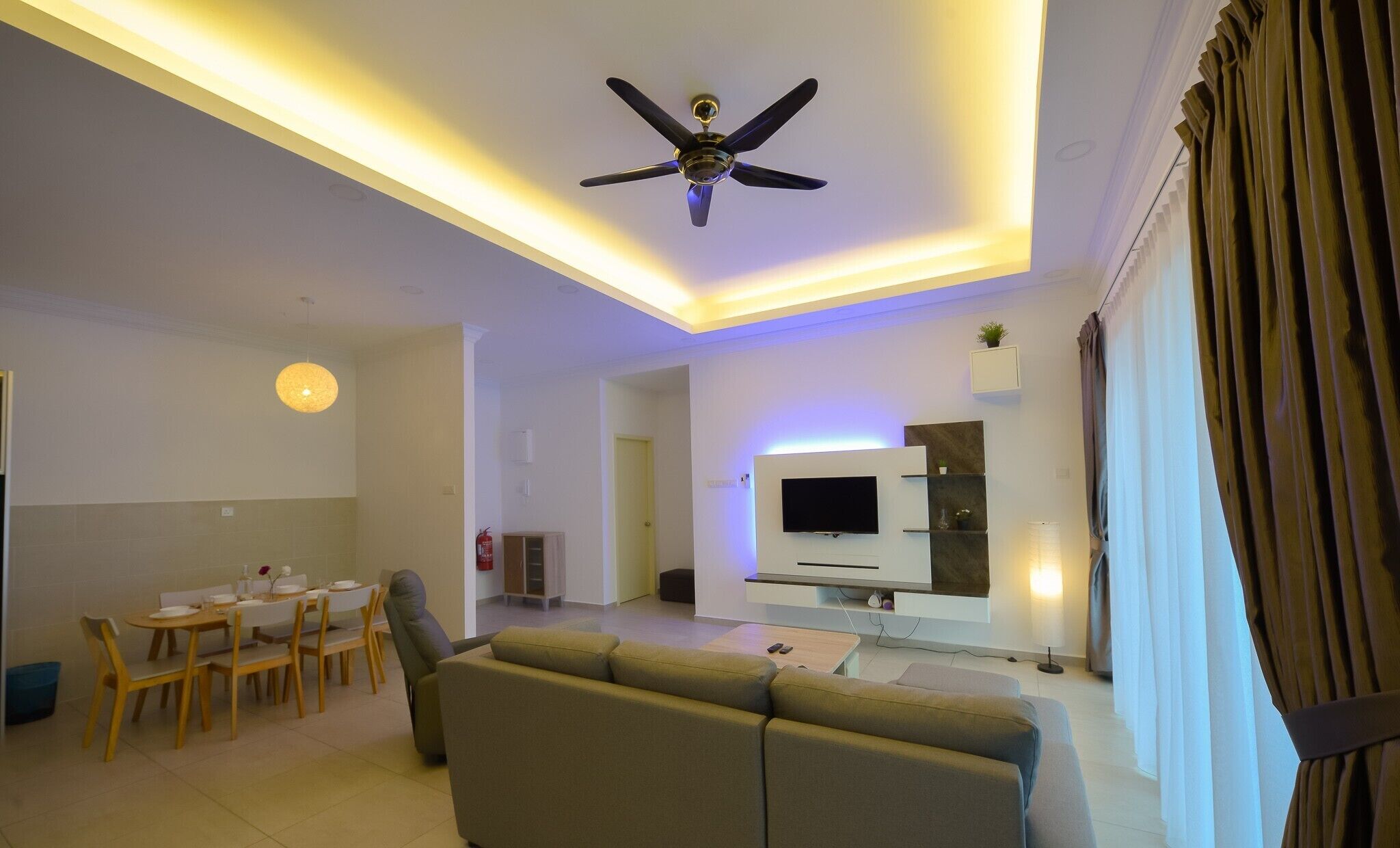 A modern living room with a ceiling fan, a dining area with a table and chairs, a sofa, and a TV mounted on the wall.