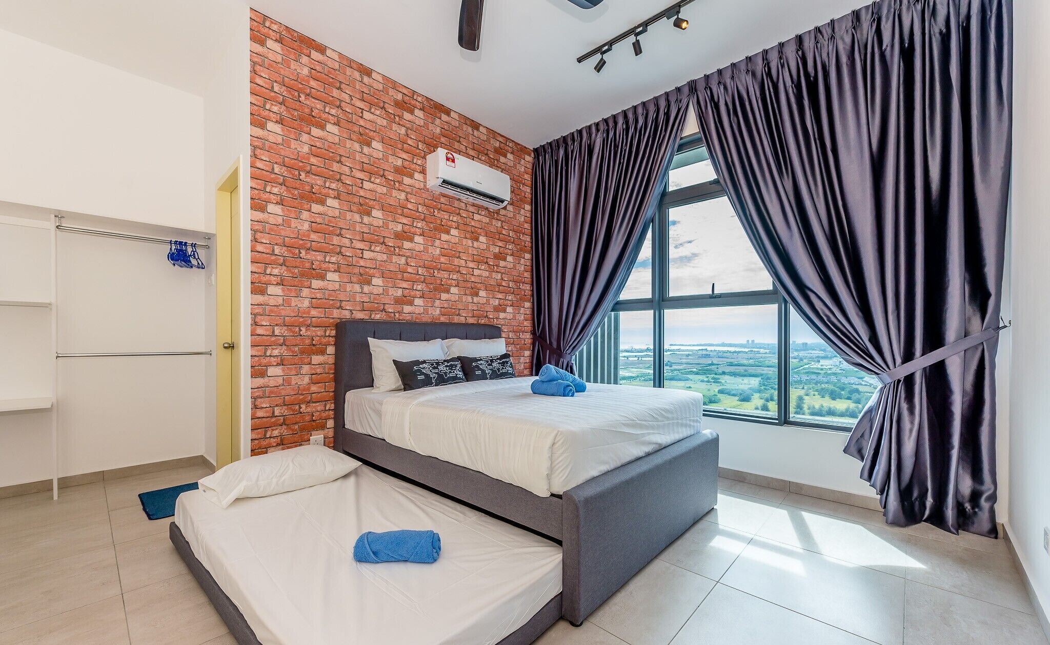 A modern hotel room with a brick accent wall, a large window with curtains, a bed with pillows, and a flat-screen TV.