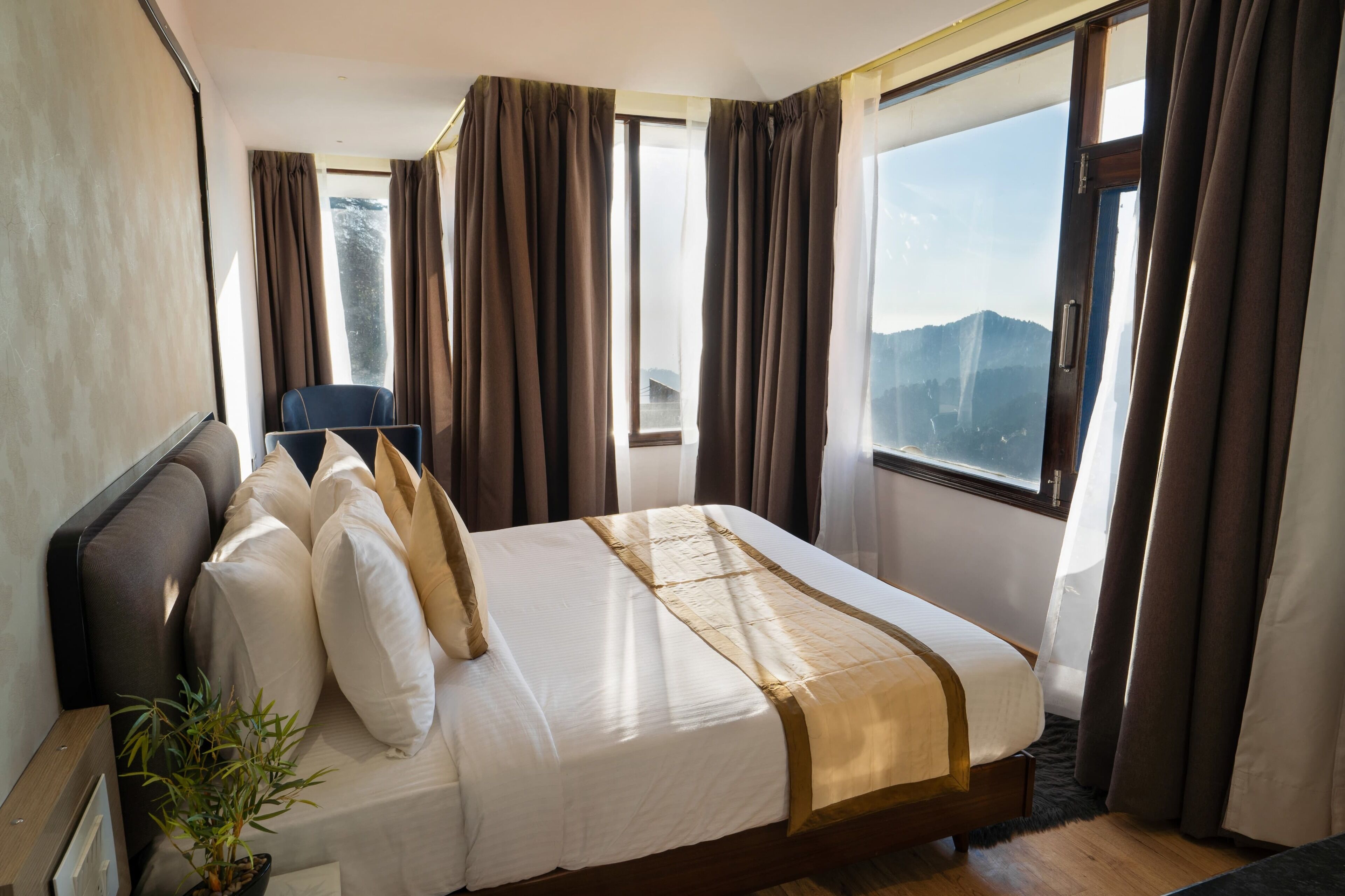 Quarto de hotel com cama grande, uma cadeira, planta em vaso e vista para montanhas pela janela.