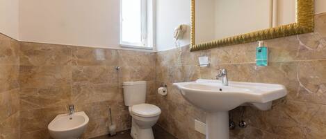 Baño con bidé, inodoro y lavamanos, todo con paredes de mármol y un espejo grande.