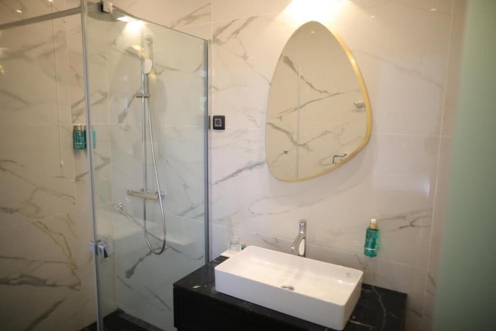 Een moderne badkamer met een douchecabine van glas, een wit wastafelmeubel en een marmeren wand met een spiegel met gouden lijst.
