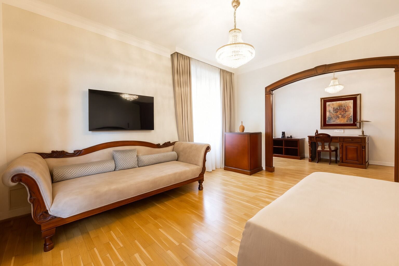 A hotel room with a wooden floor, a sofa, a bed, a desk, and a television mounted on the wall.