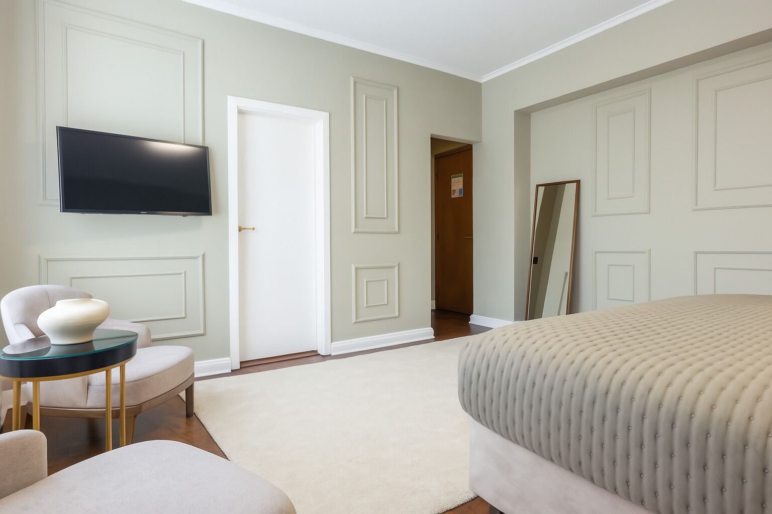 A bedroom with a bed, a TV mounted on the wall, a chair, a small table, and a door leading to another room.