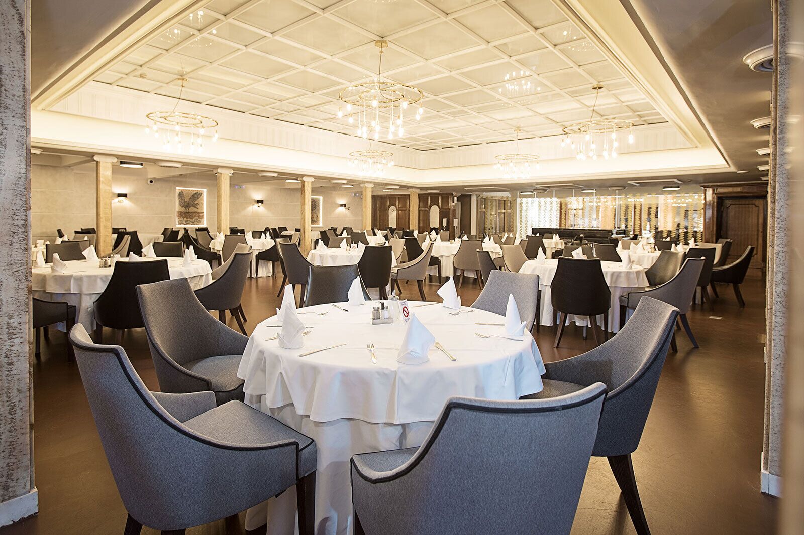 A large banquet hall with round tables and grey chairs, featuring a high ceiling with chandeliers and recessed lighting.