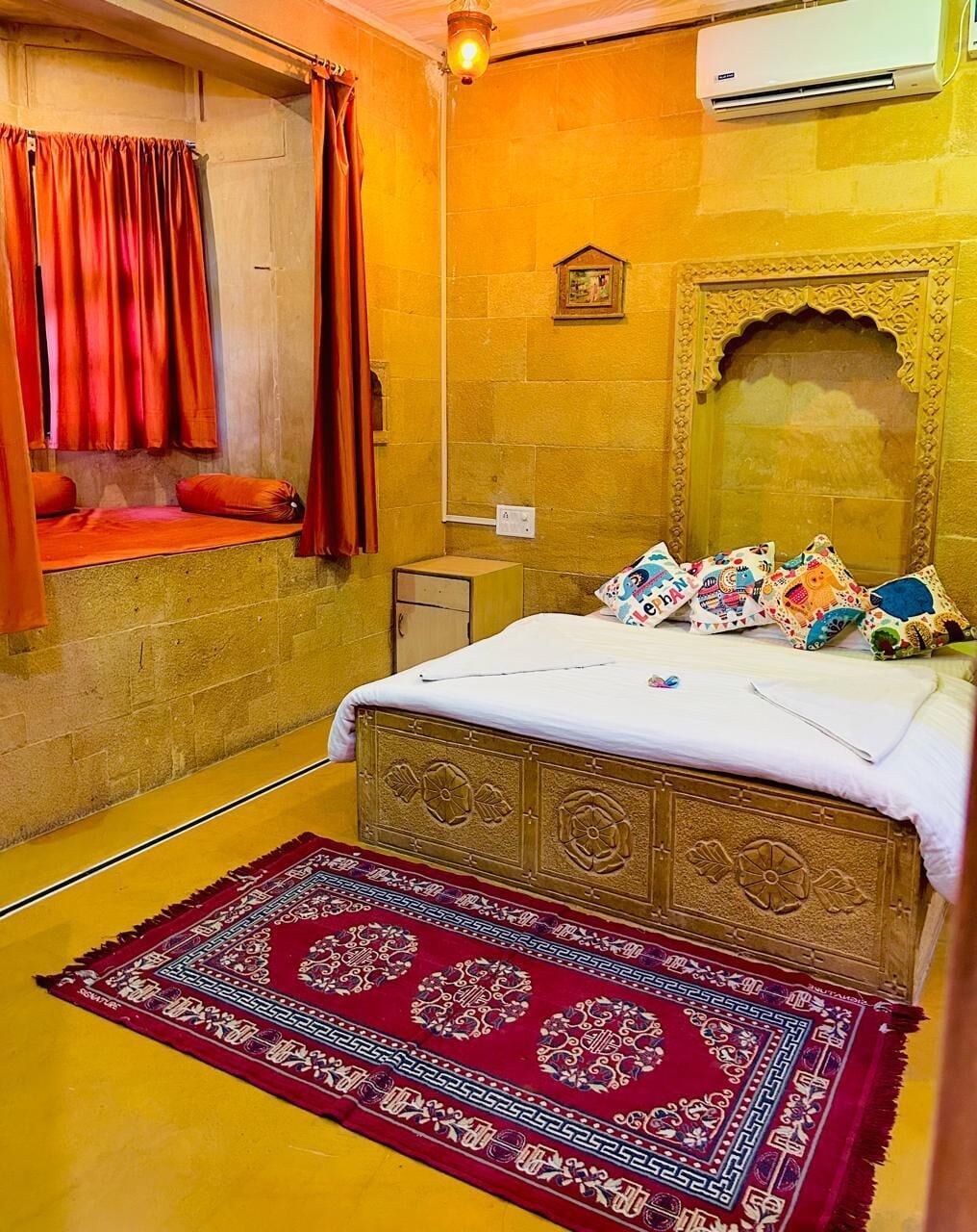 A room with a bed, a red patterned rug, a wall-mounted air conditioner, and a window with red curtains.