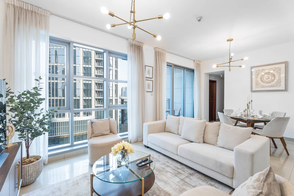A modern living room with a large window, a sofa, a coffee table, and a dining area with a chandelier.