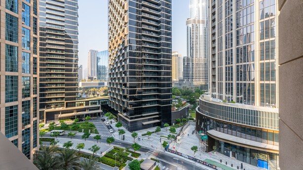 Una zona urbana moderna con edificios altos, una plaza bien cuidada con árboles y un cielo despejado.