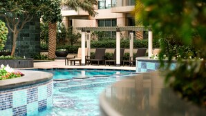 Una piscina con sillones de descanso y una pérgola, rodeada de follaje y un edificio al fondo.