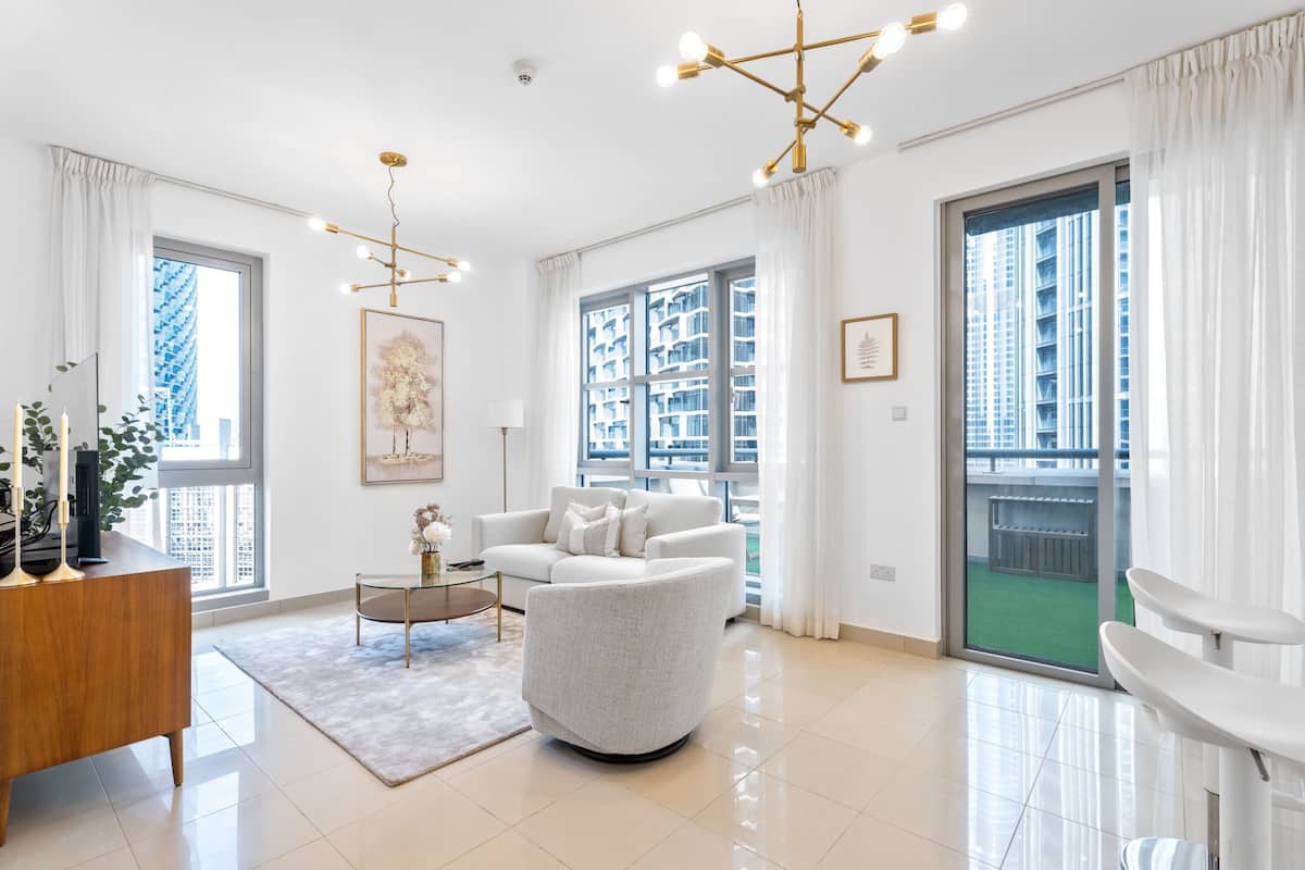 A modern living room with a white sofa, a coffee table, and a chandelier. There is a large window with a view of buildings.