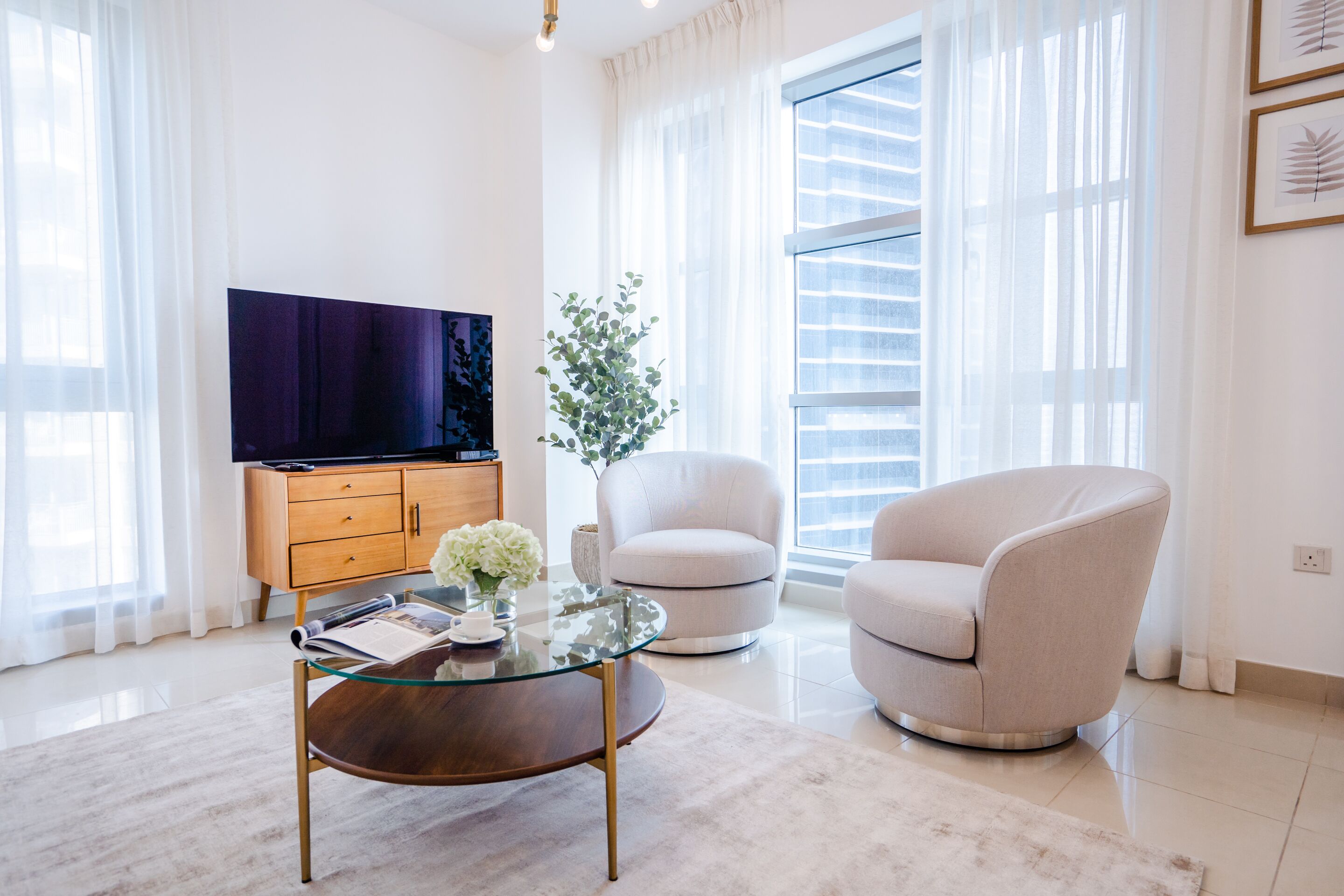 A modern living room with a glass coffee table, a beige armchair, a wooden cabinet, and a flat-screen TV.
