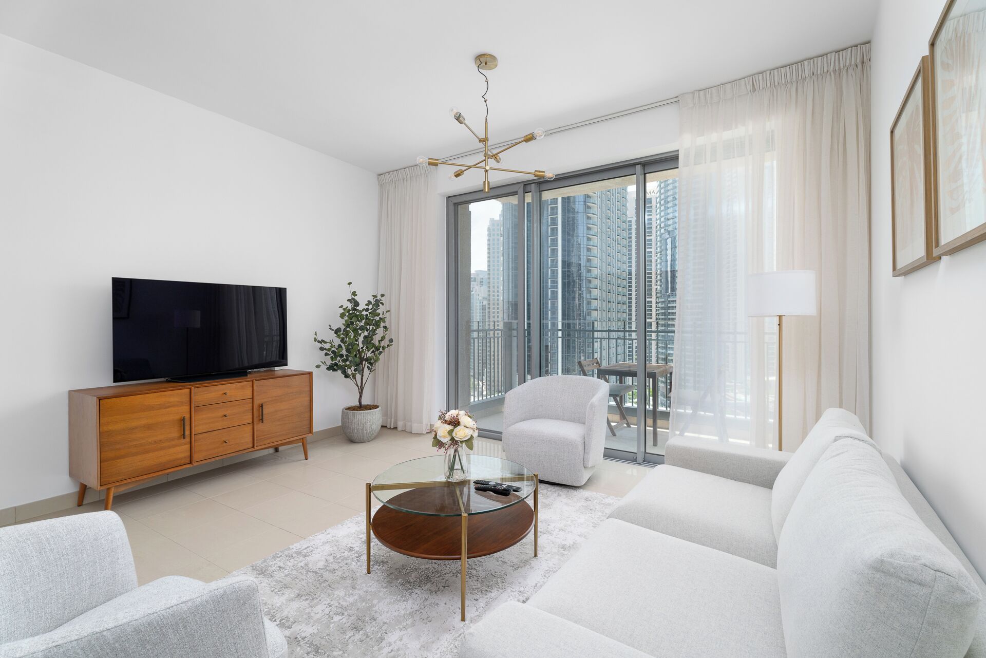A modern living room with a flat-screen TV, a wooden cabinet, a glass coffee table, a grey sofa, and a large window with a city view.
