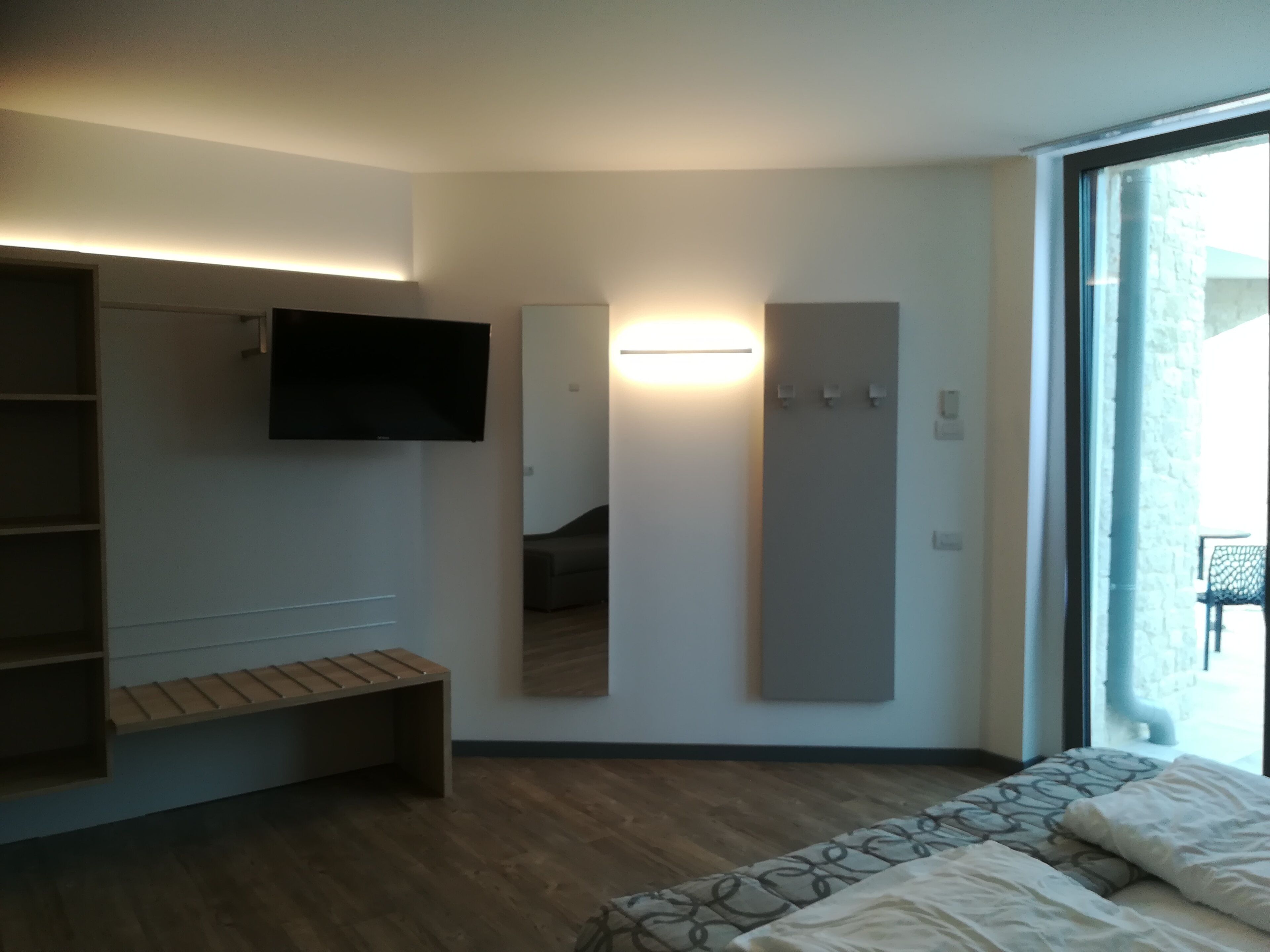 Ein modernes Hotelzimmer mit einem großen Bett, einem flachbildfernseher an der Wand, einer Bank und einer Schiebetür, die auf einen Balkon führt.