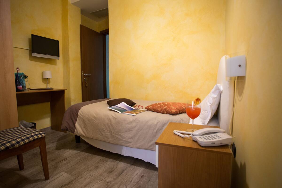 Ein Hotelzimmer mit einem Bett, einem Nachttisch mit Telefon, einer Lampe, einem an der Wand befestigten Fernseher und einer Tür zu einem weiteren Raum.
