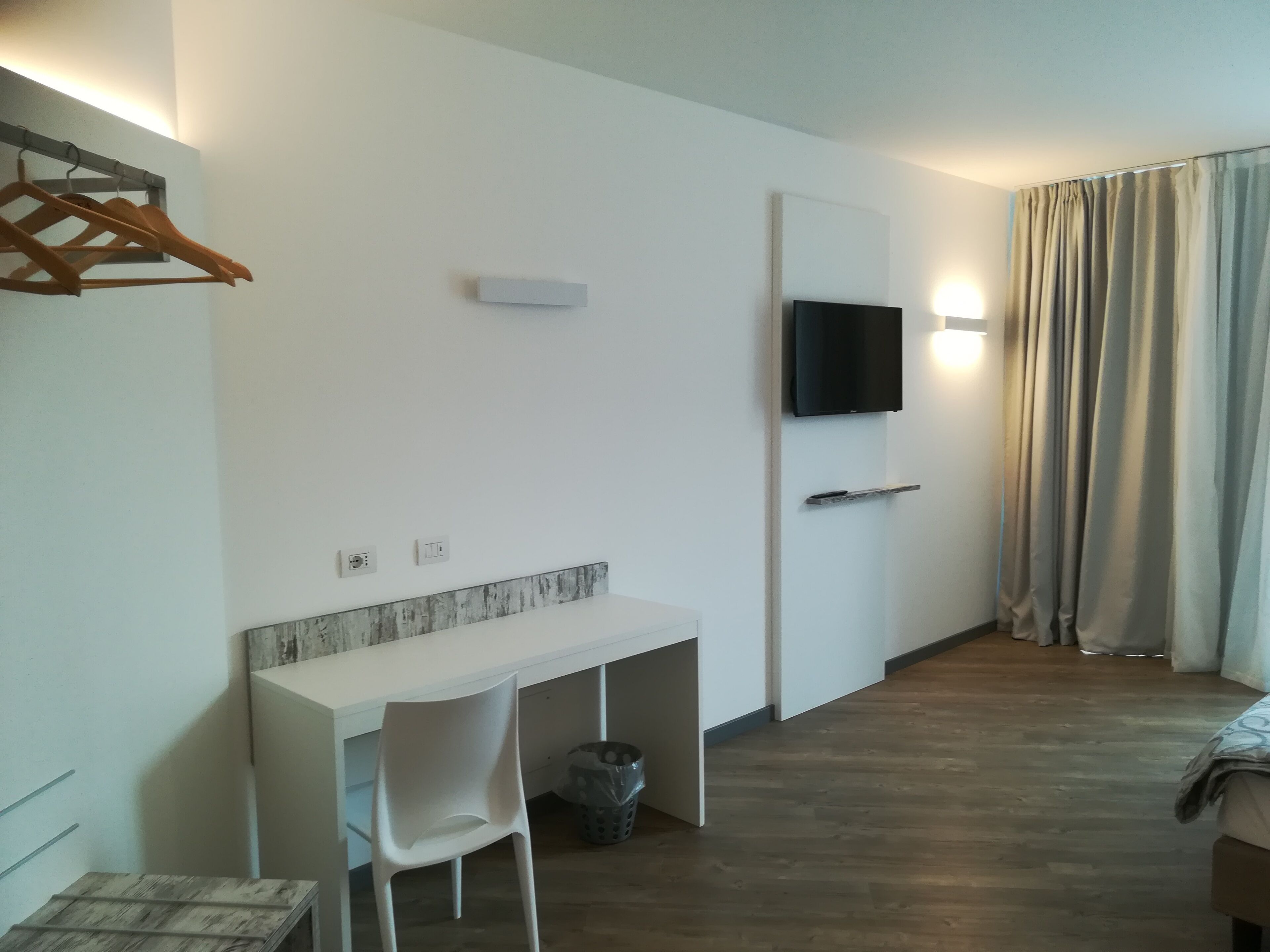 Ein modernes Hotelzimmer mit weißem Schreibtisch, einem Sessel, einem an der Wand befestigten Fernseher und einem fensterhohen Vorhang.