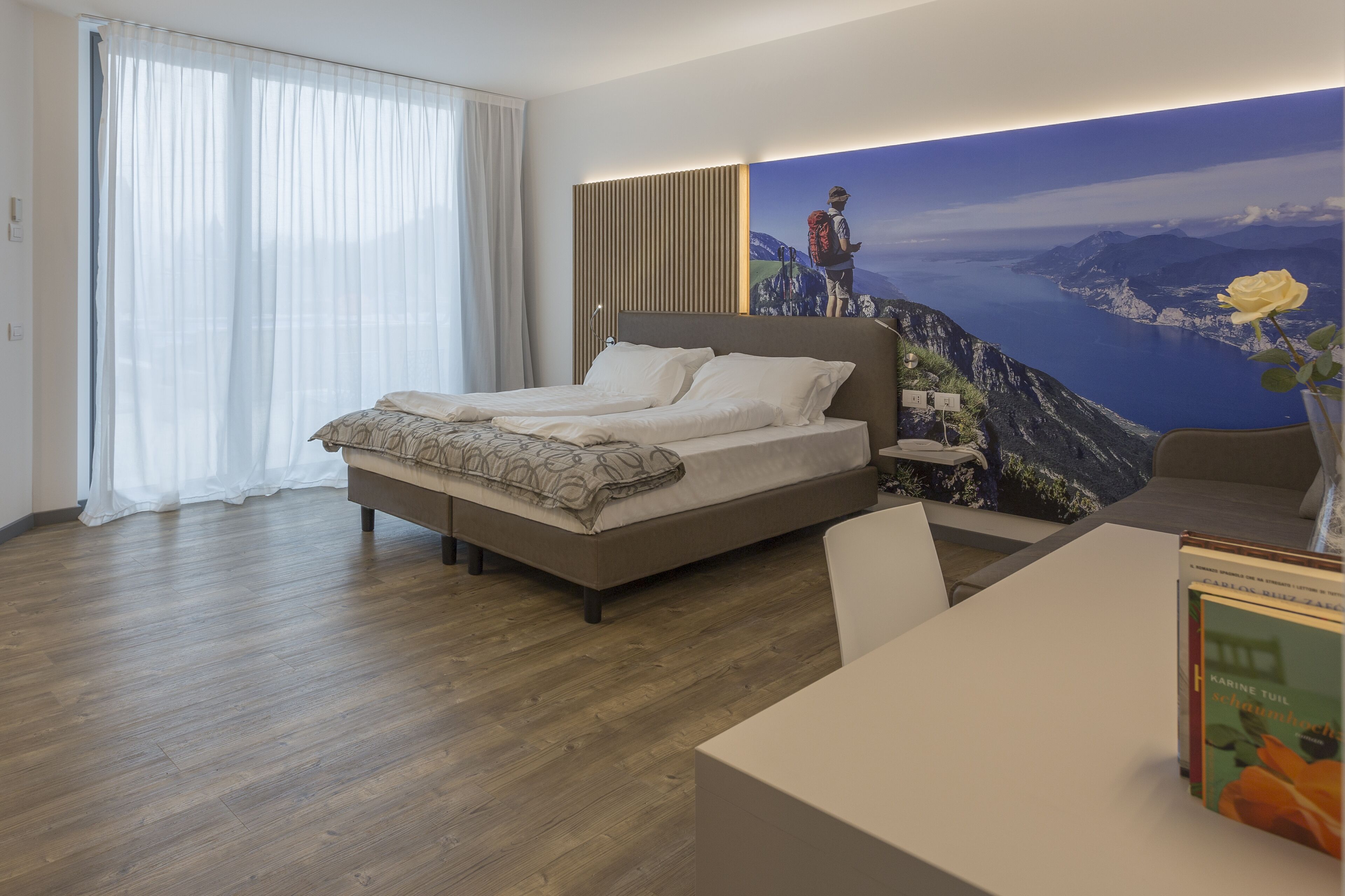 A modern hotel room with a large bed, a view of a mountainous landscape, and a desk with a vase of flowers.