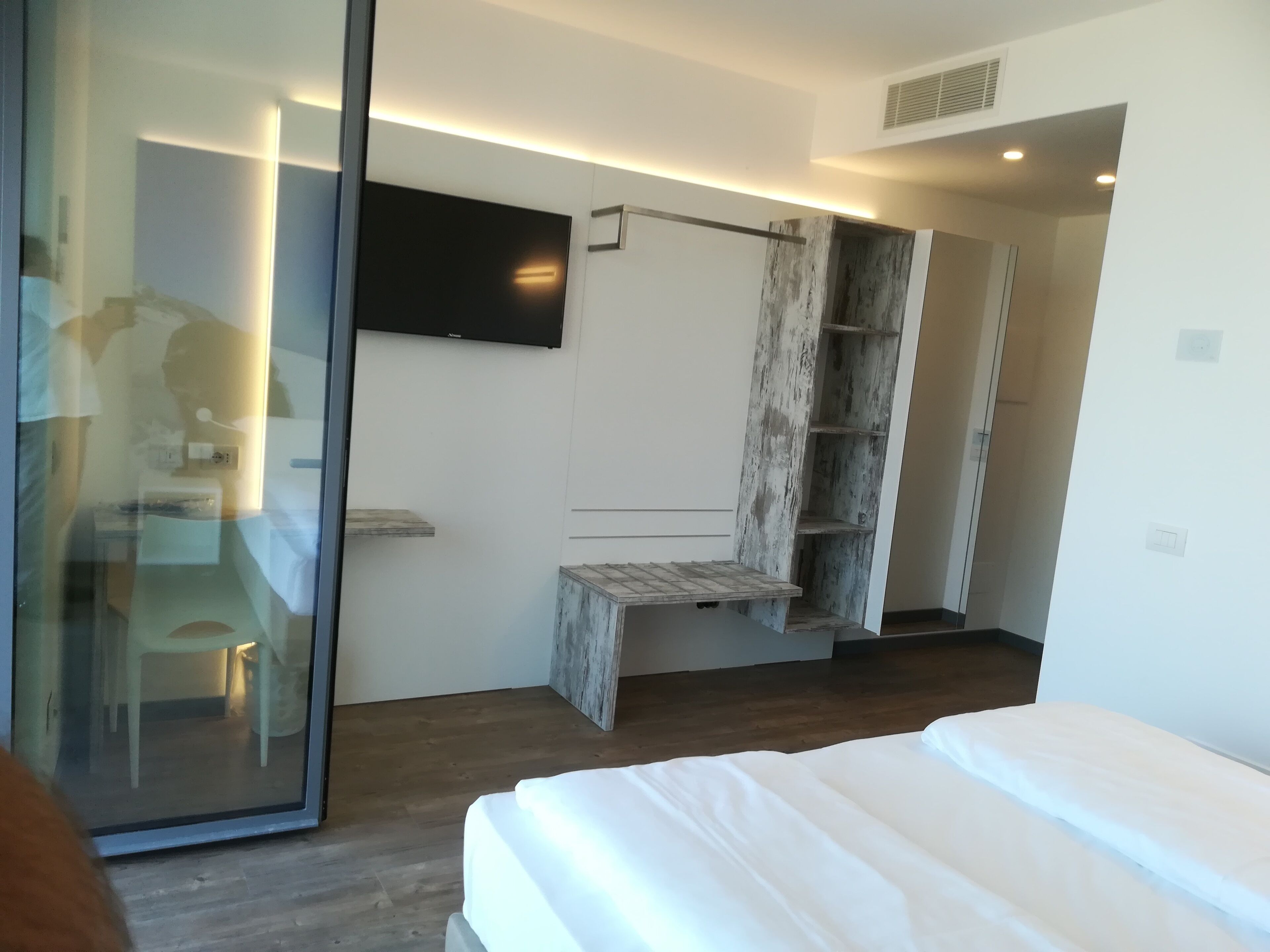A modern hotel room with a large bed, a flat-screen TV mounted on the wall, and a wooden shelf unit.