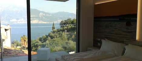 A modern bedroom with a large bed, a view of a coastal landscape, and a balcony with a white table.