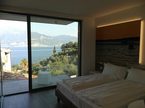 A modern bedroom with a large bed, a view of a coastal landscape, and a balcony with a white table.